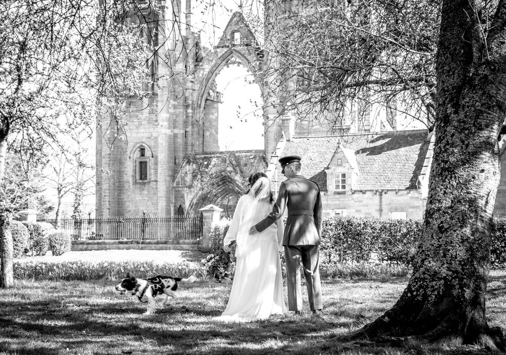 A black and white wedding photograph showing a couple and their dog beside a gothic cathedral structure.