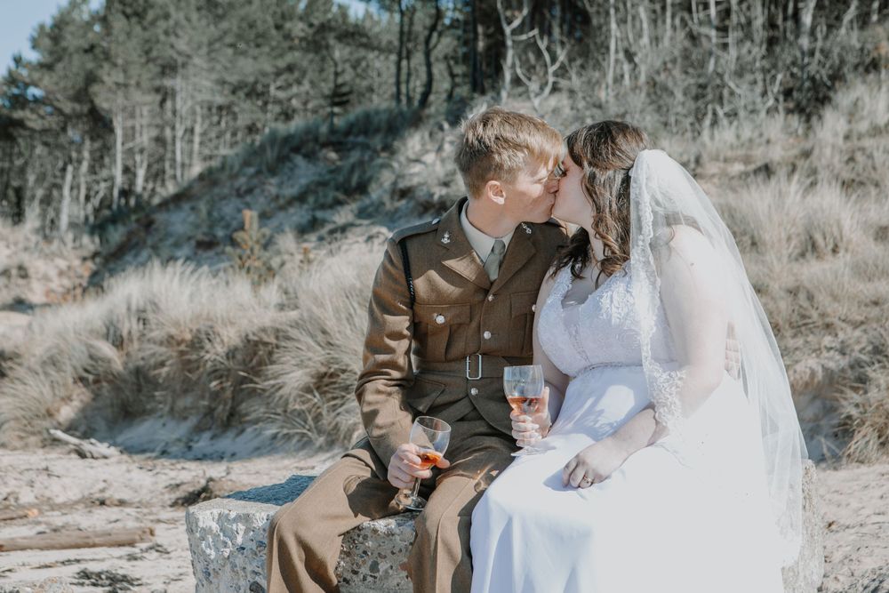 A romantic couple in wedding attire sits together on rocks with mountains and trees in the background.