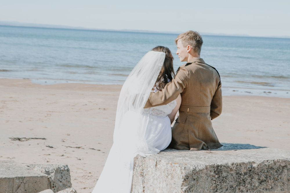 A couple wearing wedding attire sits together on a concrete barrier at the beach looking out at the ocean.