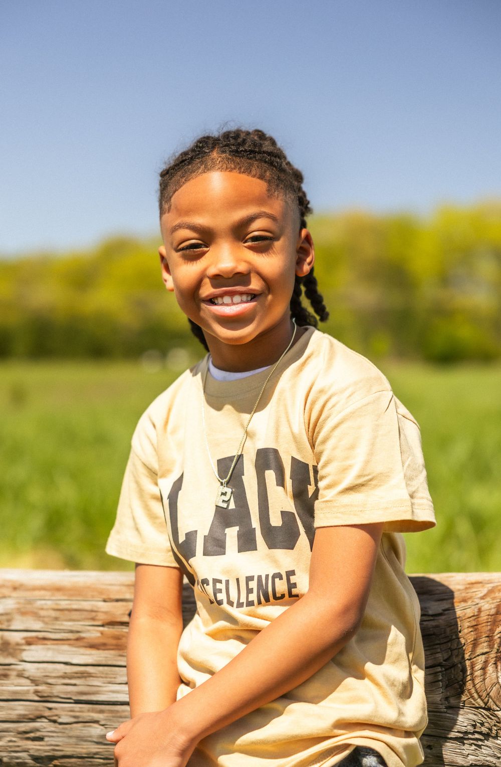 Smiling youth in a beige 'Black Excellence' t-shirt sits outdoors on a sunny day with green foliage in background.