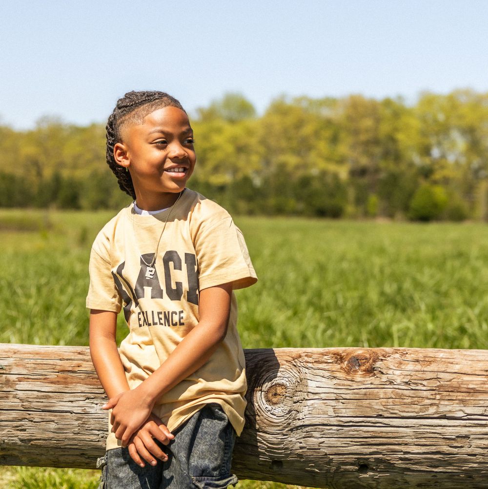 Smiling youth in a beige t-shirt sits on rustic wooden fence with green countryside landscape in background.
