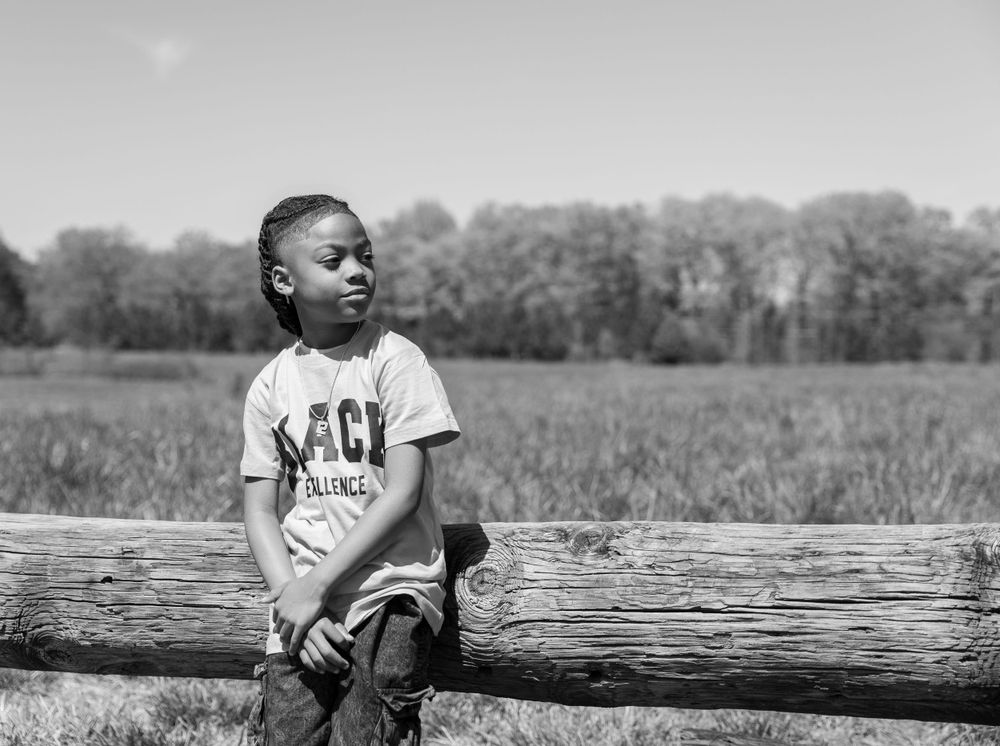 Black and white photo of a child in a t-shirt sitting on a wooden log fence in a rural field.