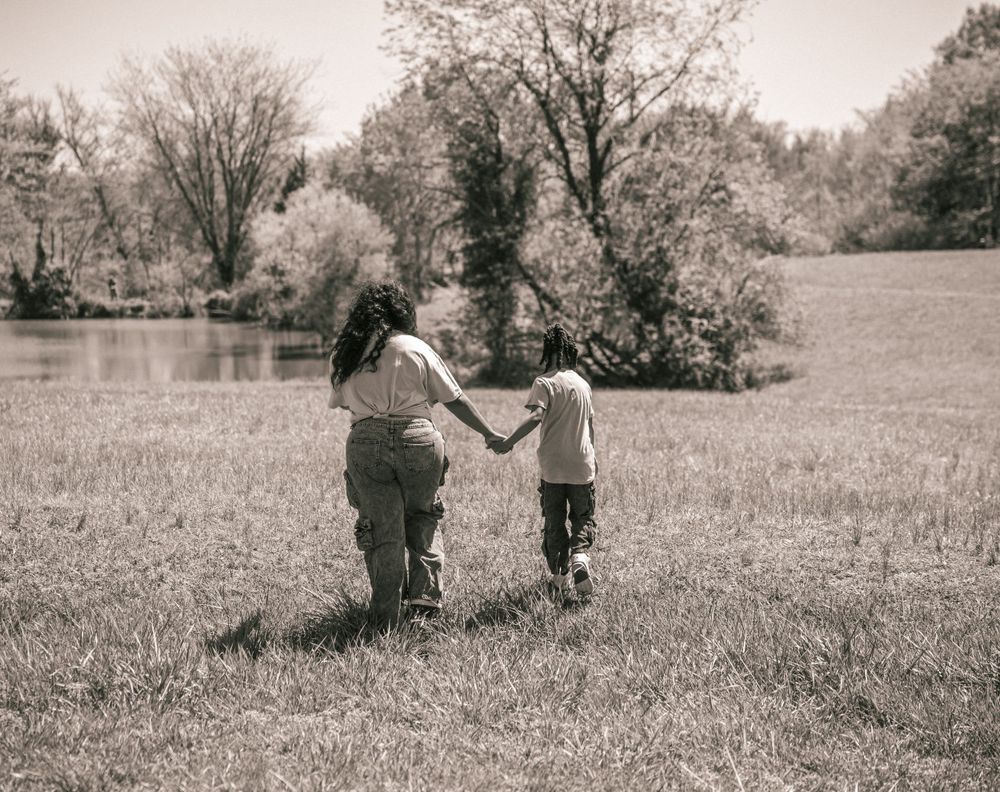 Two children holding hands while walking through a grassy field near a lake in black and white.