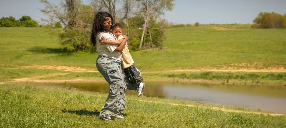 Person in camouflage pants stands fishing by a pond on a sunny spring day with green grass and trees in background.