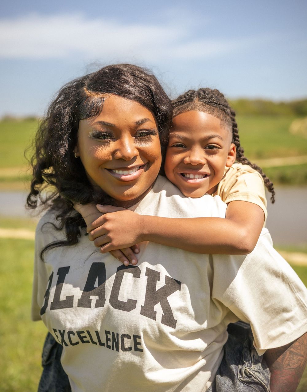 Two people embracing and smiling outdoors on a sunny day, with one wearing a 'BLACK EXCELLENCE' t-shirt.