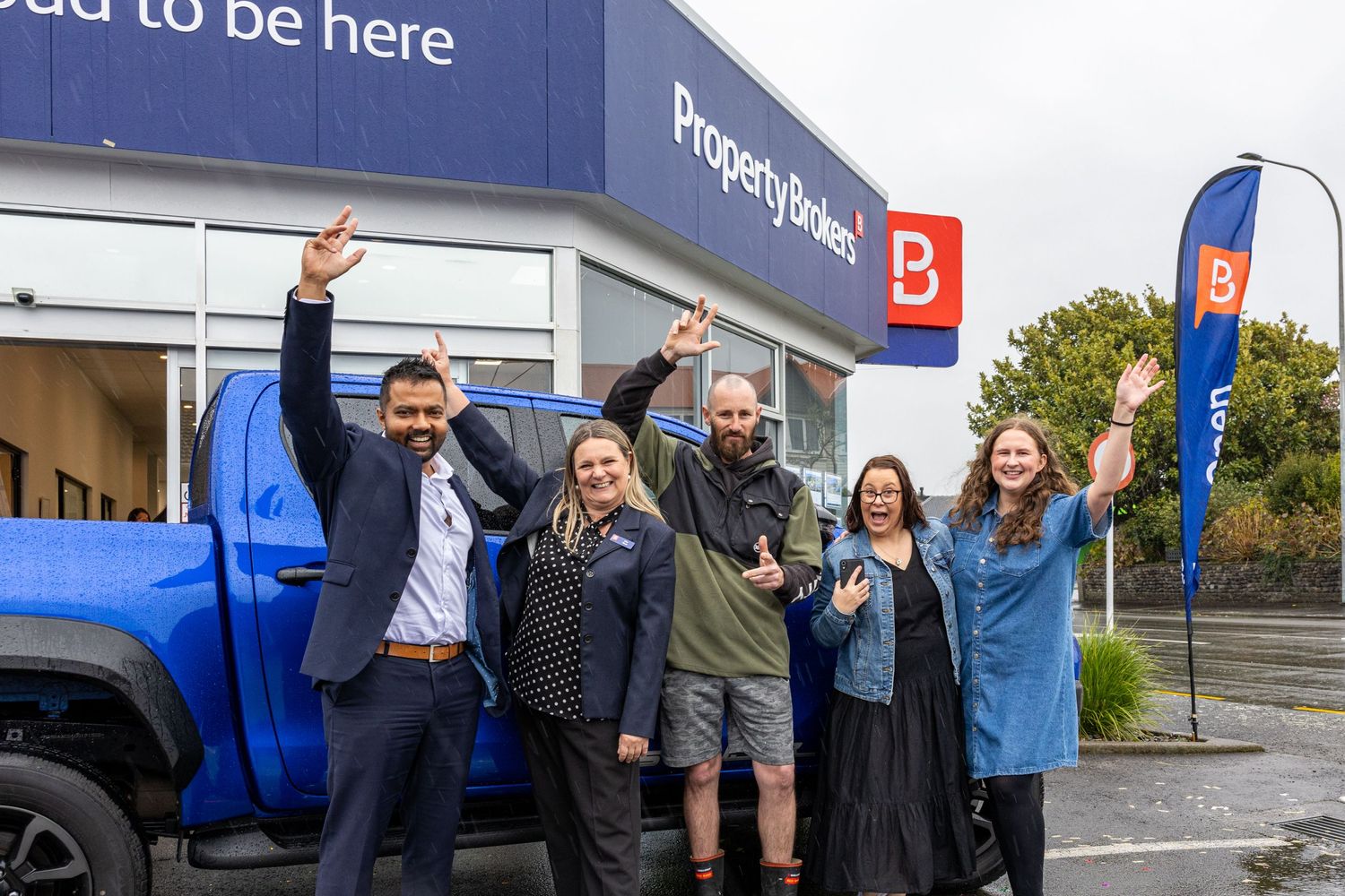 A group of excited people celebrating outside a retail storefront with blue signage and vehicle visible.