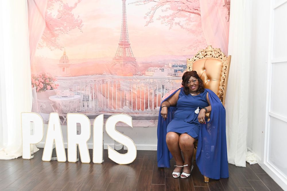A person in a royal blue dress poses against a white wall with pink sunset-colored artwork in the background.