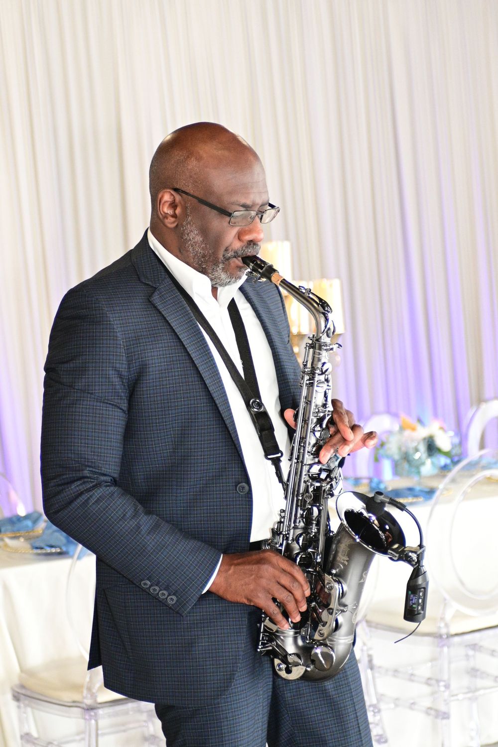Saxophone player in navy suit performs at elegant event with white drapes and decor in background.