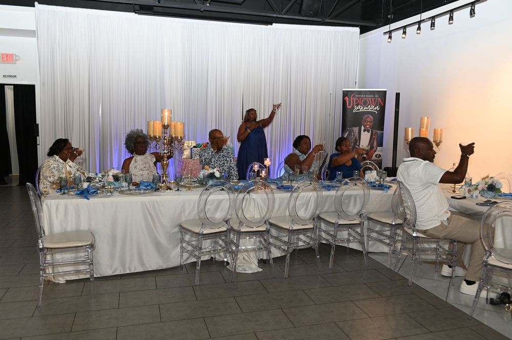 Event venue setup showing multiple white chairs and tables arranged in rows with white draping and lighting elements.