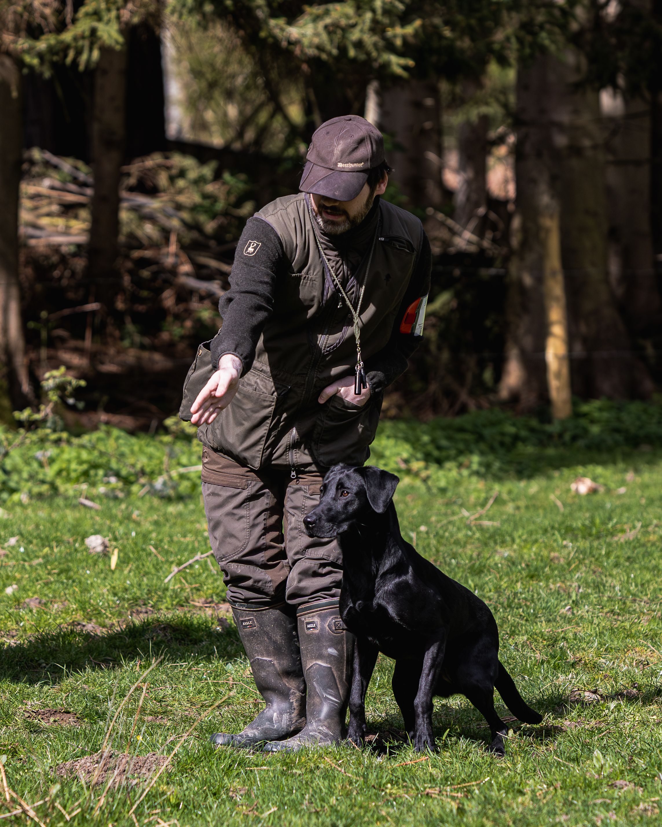 A dog trainer in dark outdoor gear and rubber boots works with a black Labrador Retriever on green grass in a forest.