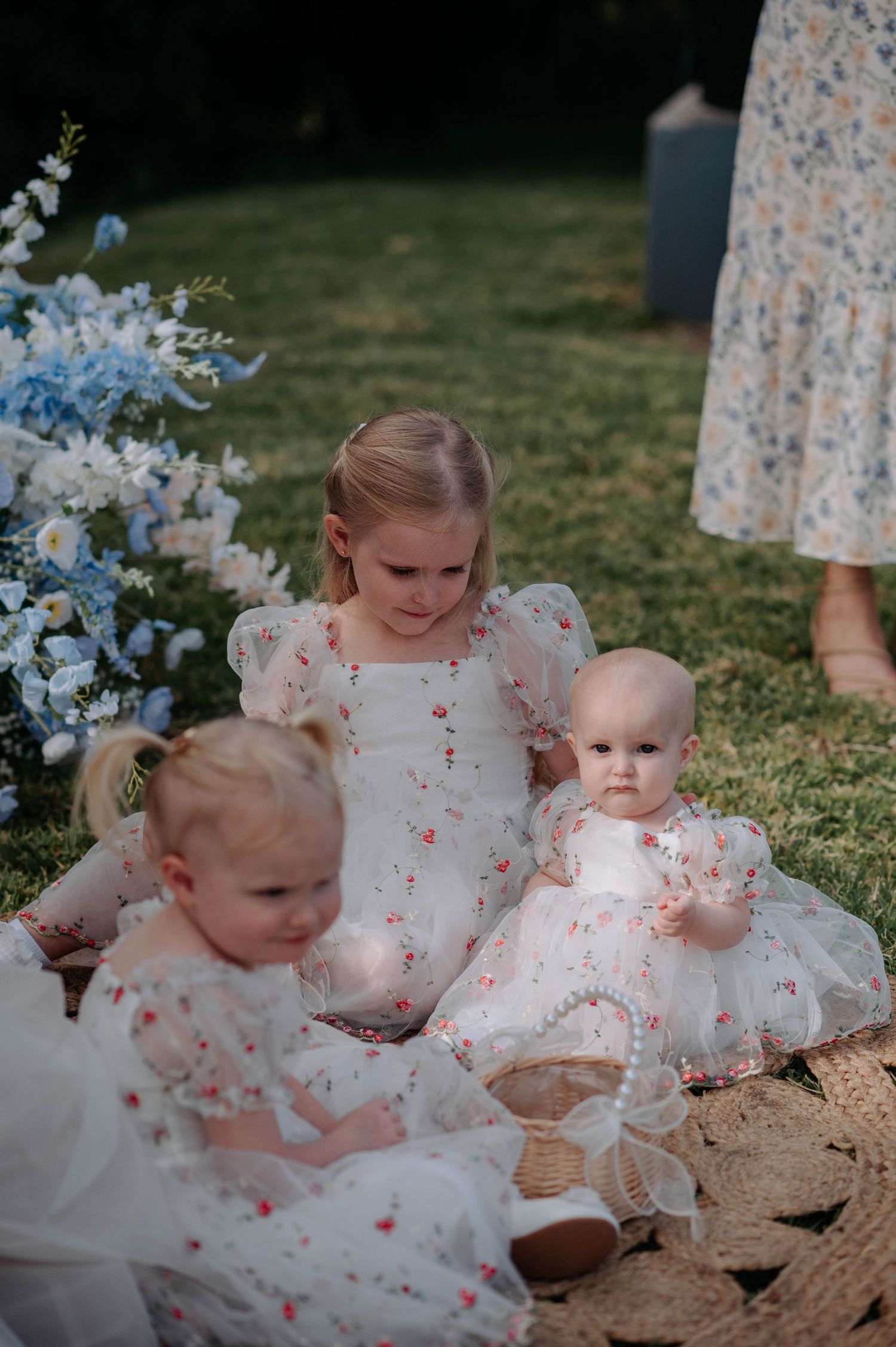 Young children in matching white floral dresses sit together on a blanket near blue flowers.