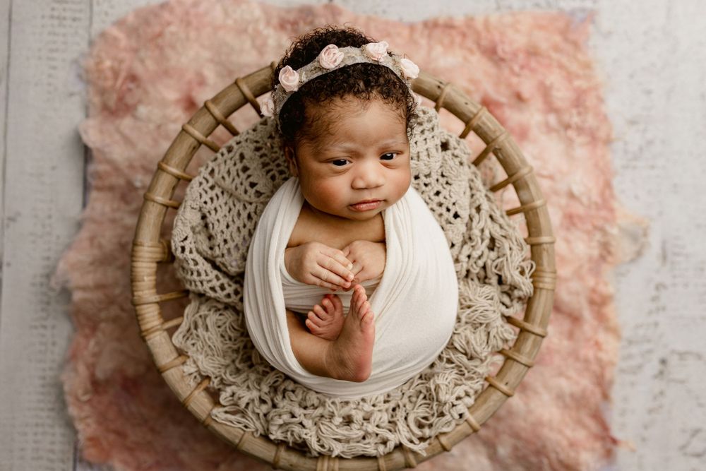 Newborn baby wrapped in white swaddle lying in round woven basket with lace details on pink fluffy backdrop.