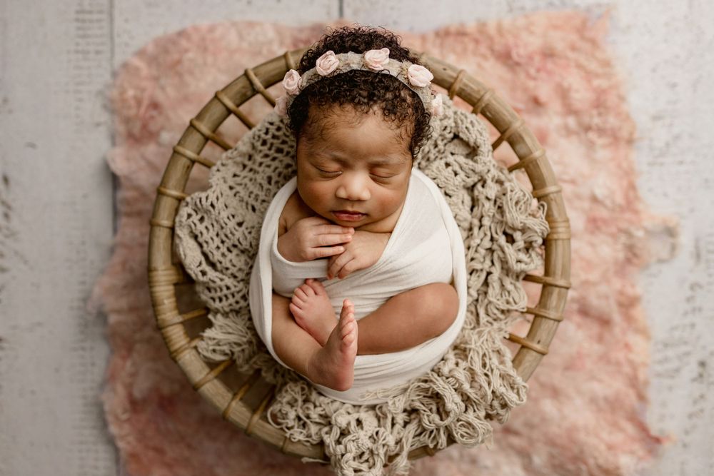 Newborn baby sleeping peacefully in a round wicker basket with cream lace and pink plush blanket.