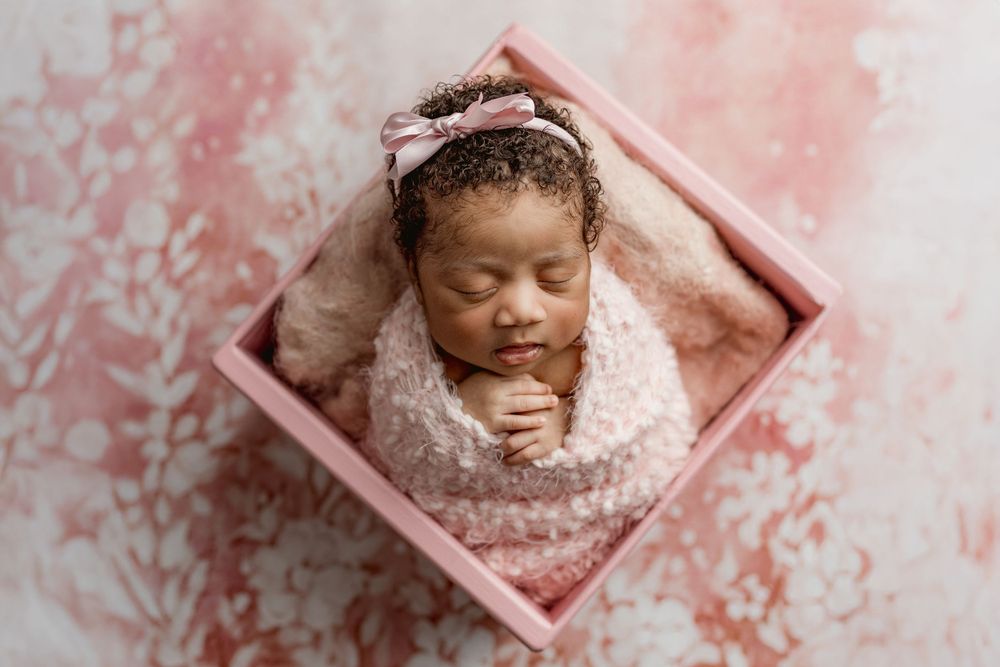 Sleeping newborn wrapped in soft fabric resting in pink box with textured pink background.