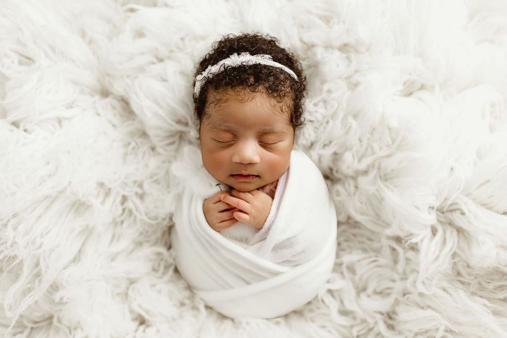 Sleeping baby wrapped in white swaddling cloth on soft white fur blanket with delicate headband.