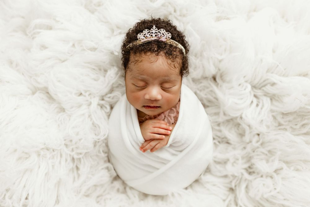 A sleeping newborn wrapped in white fabric with a sparkly headpiece lies on a plush white rug.