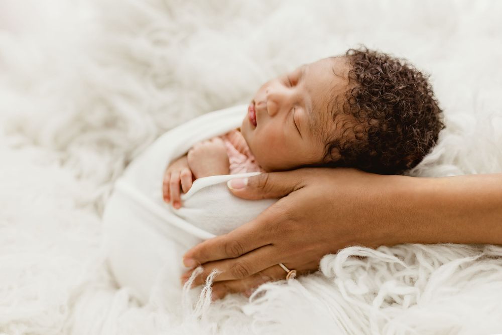 Peaceful sleeping newborn baby rests on soft white fluffy blanket with caring hands providing gentle support.