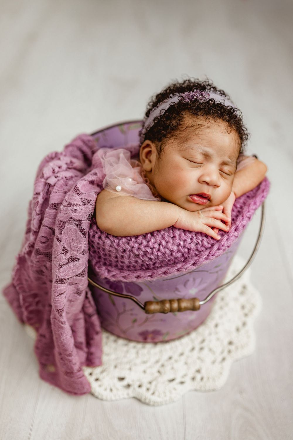 Sleeping newborn wrapped in purple knitted blanket resting in a vintage wooden bucket on white lace doily.