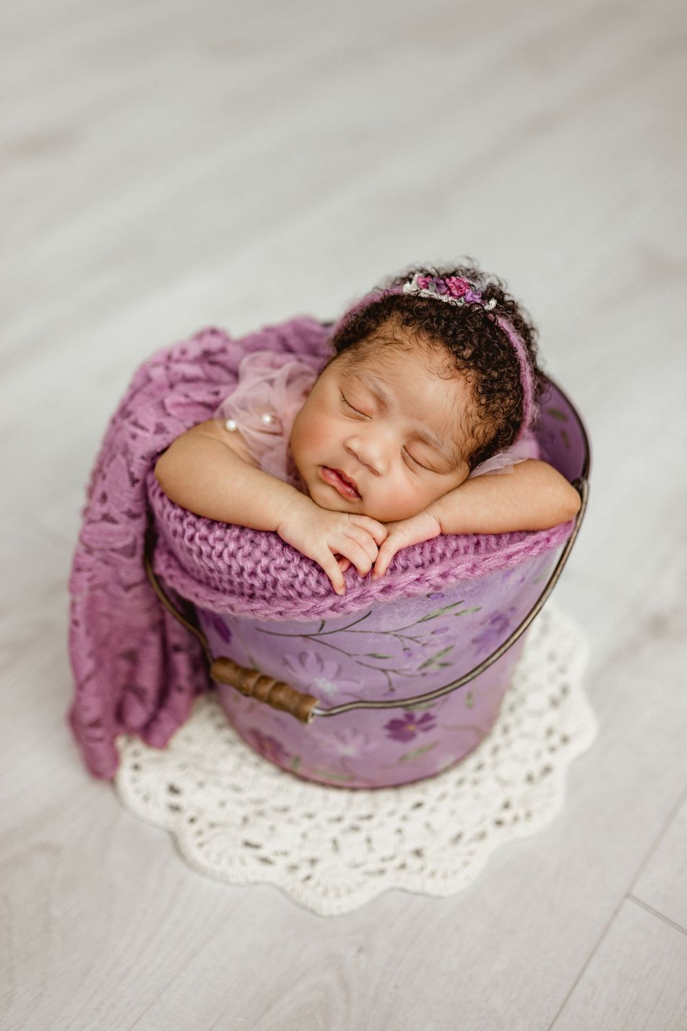 A sleeping newborn wrapped in purple knit blanket rests peacefully in a vintage floral bucket on lace doily.