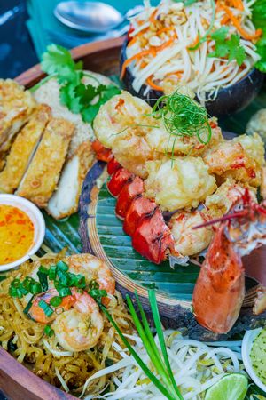 Close up of Asian fusion seafood platter with lobster tail, pad thai noodles, sushi and fresh garnishes on wooden serving tray.
