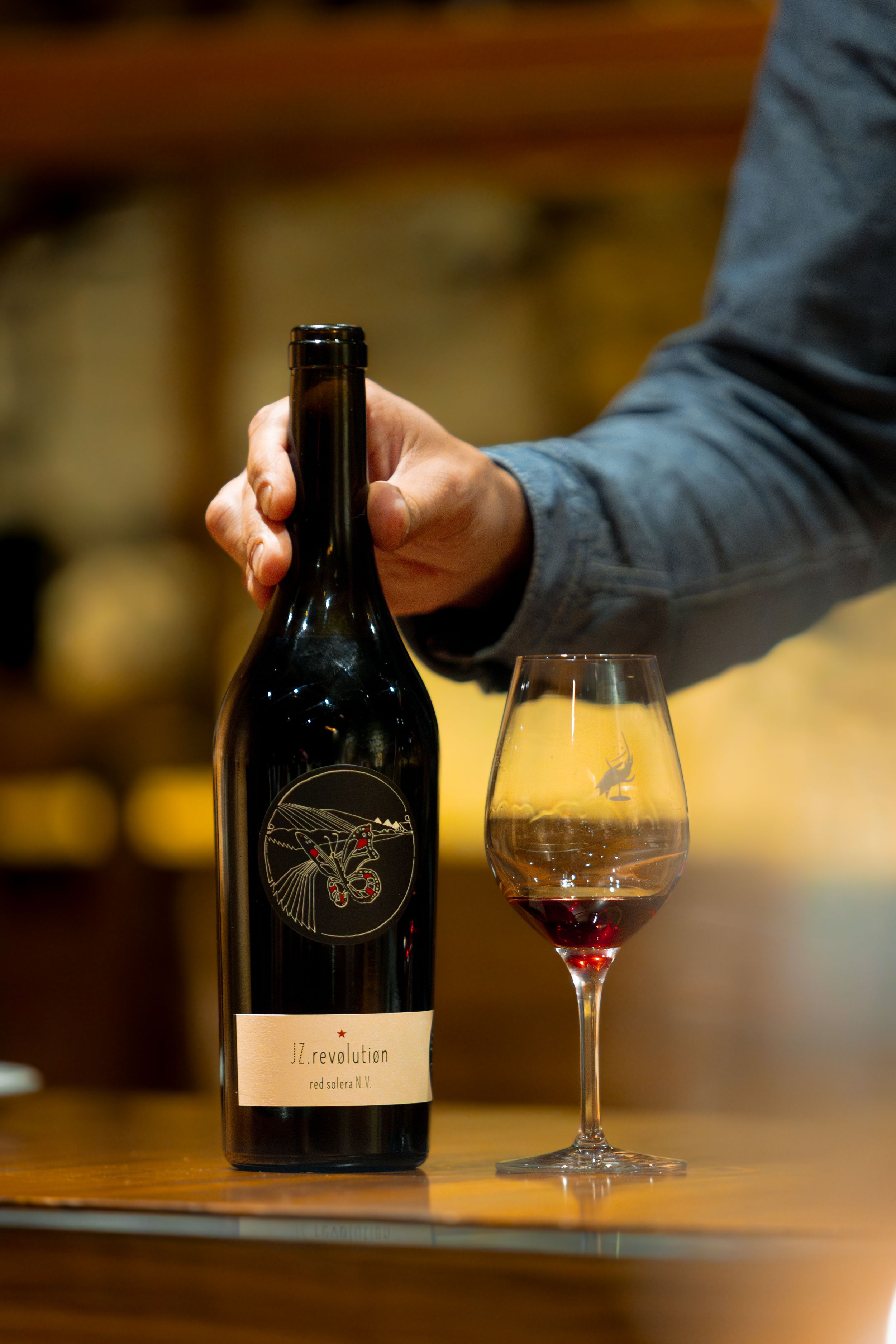 Hand holding wine bottle with elegant label next to filled wine glass on wooden surface.