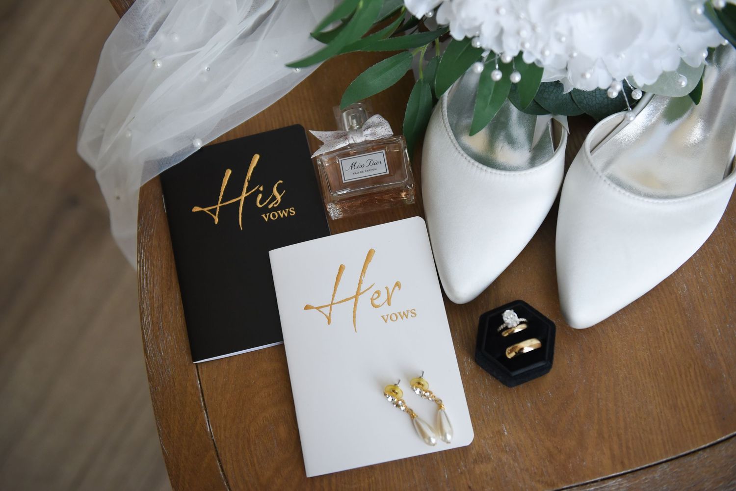 White bridal shoes, wedding vows books labeled 'His' and 'Hers', and rings displayed on wooden surface.