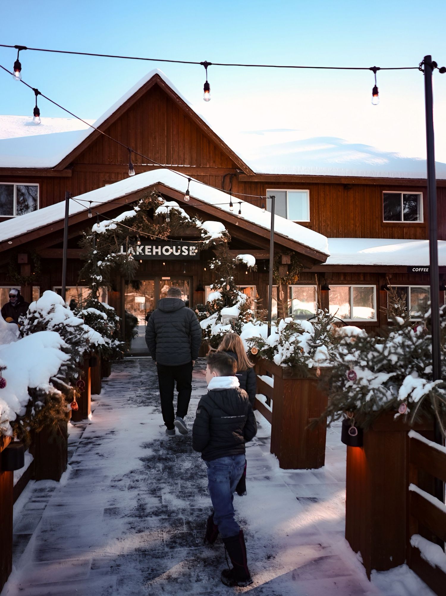 Winter walkway between wooden buildings with string lights and snow-covered landscaping.