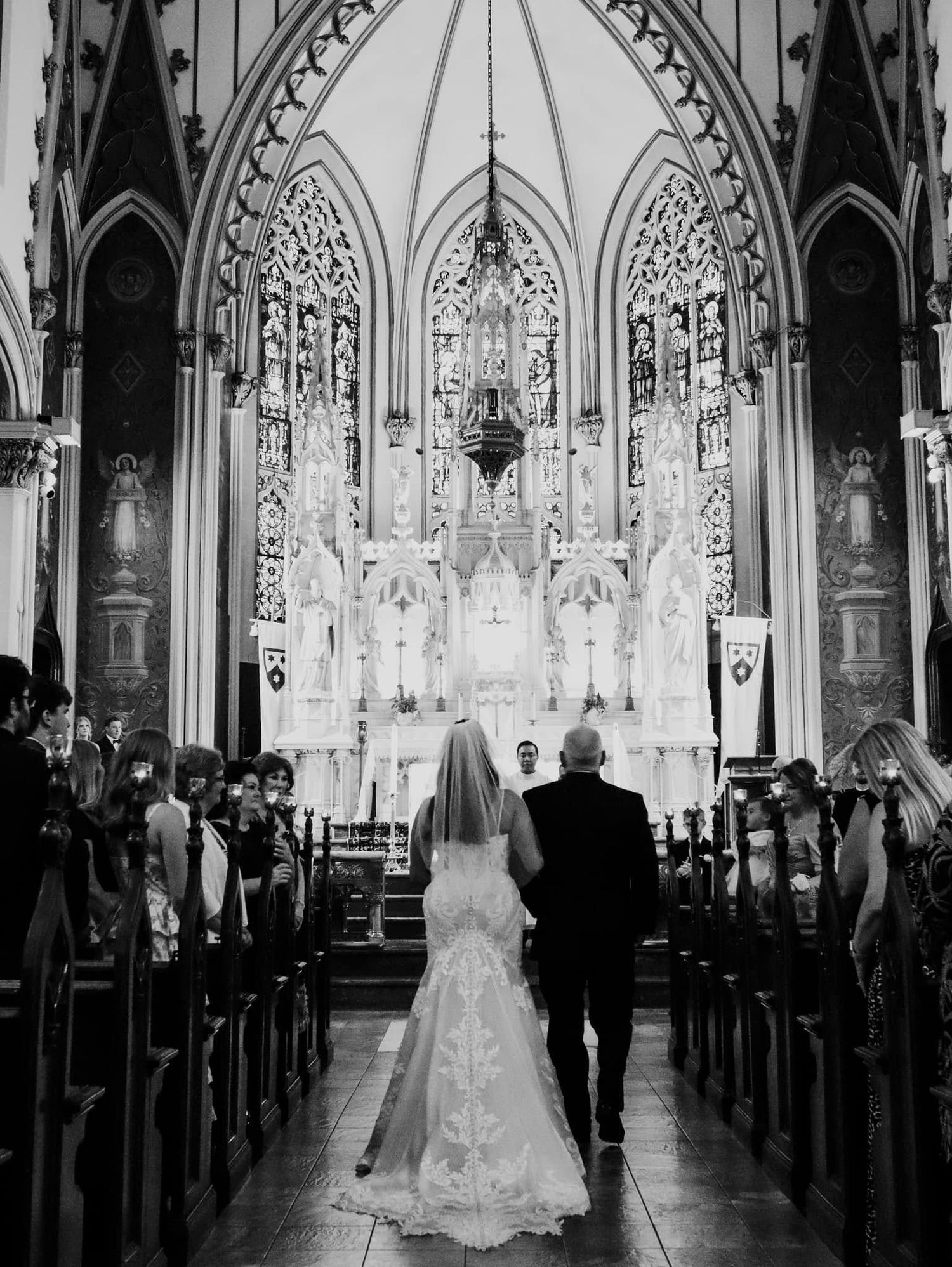 A black and white wedding ceremony photograph inside a gothic cathedral with ornate stained glass windows and vaulted arches.