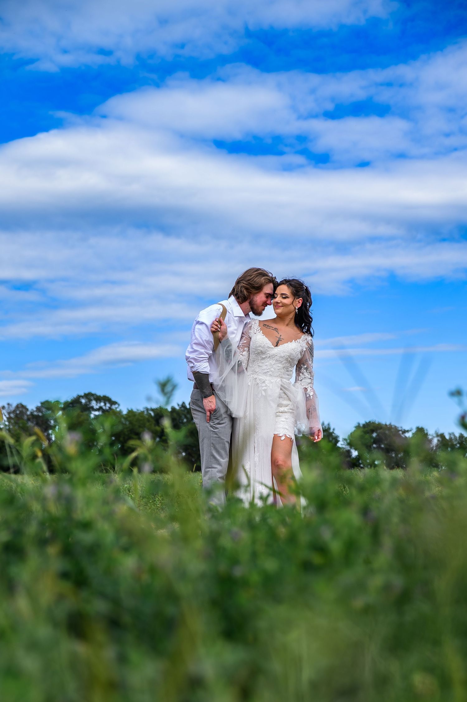 A couple shares a romantic moment in a grassy field under bright blue skies and white clouds.