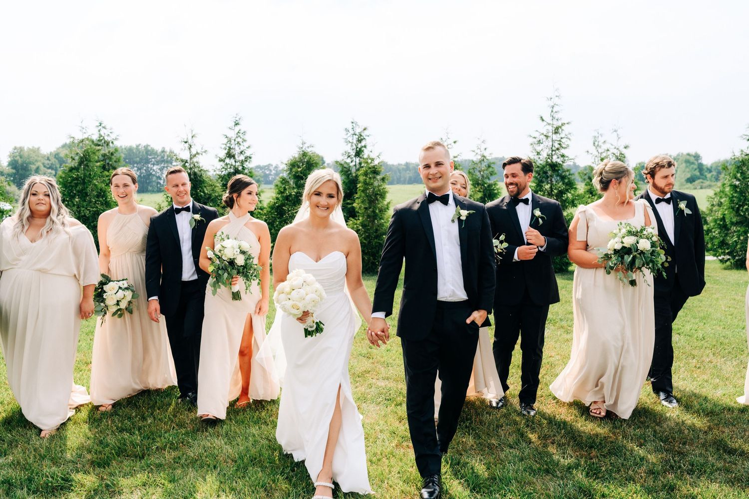 A wedding party walks and laughs together on a grassy lawn during a sunny outdoor celebration.
