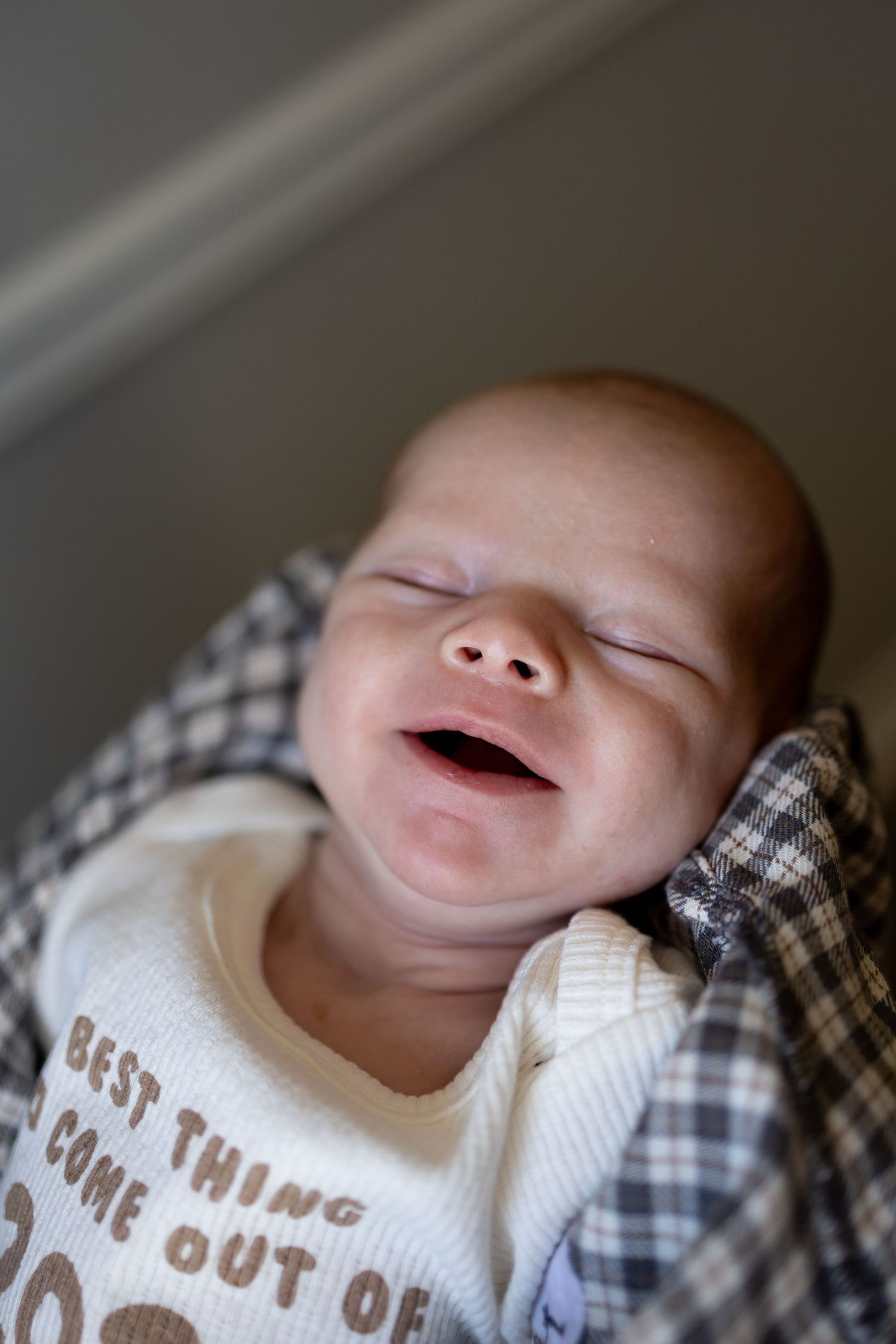 Newborn baby smiling in sleep wearing a white onesie with text, wrapped in a plaid blanket.