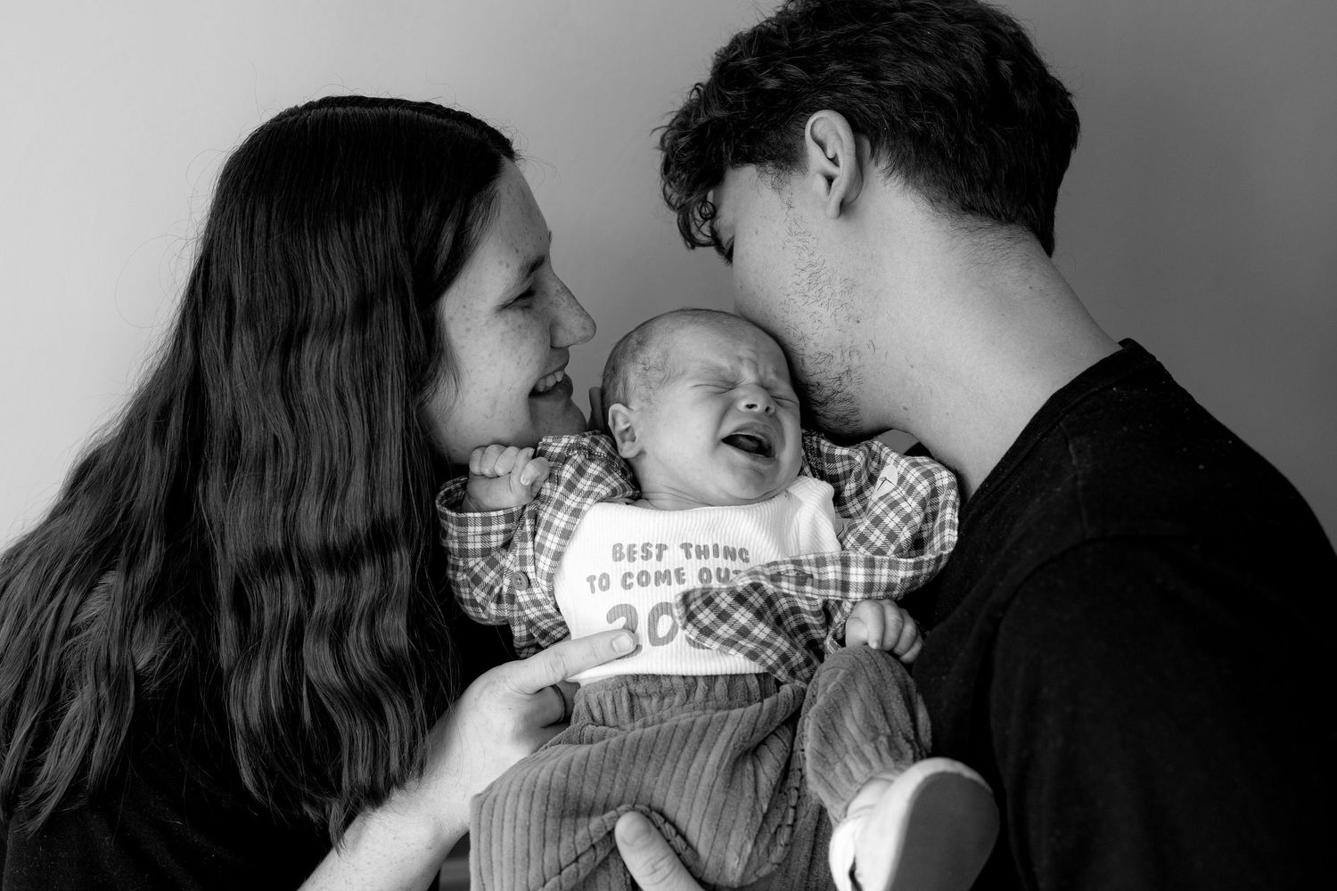 Black and white photo of new parents smiling at their sleeping newborn baby wrapped in a blanket.