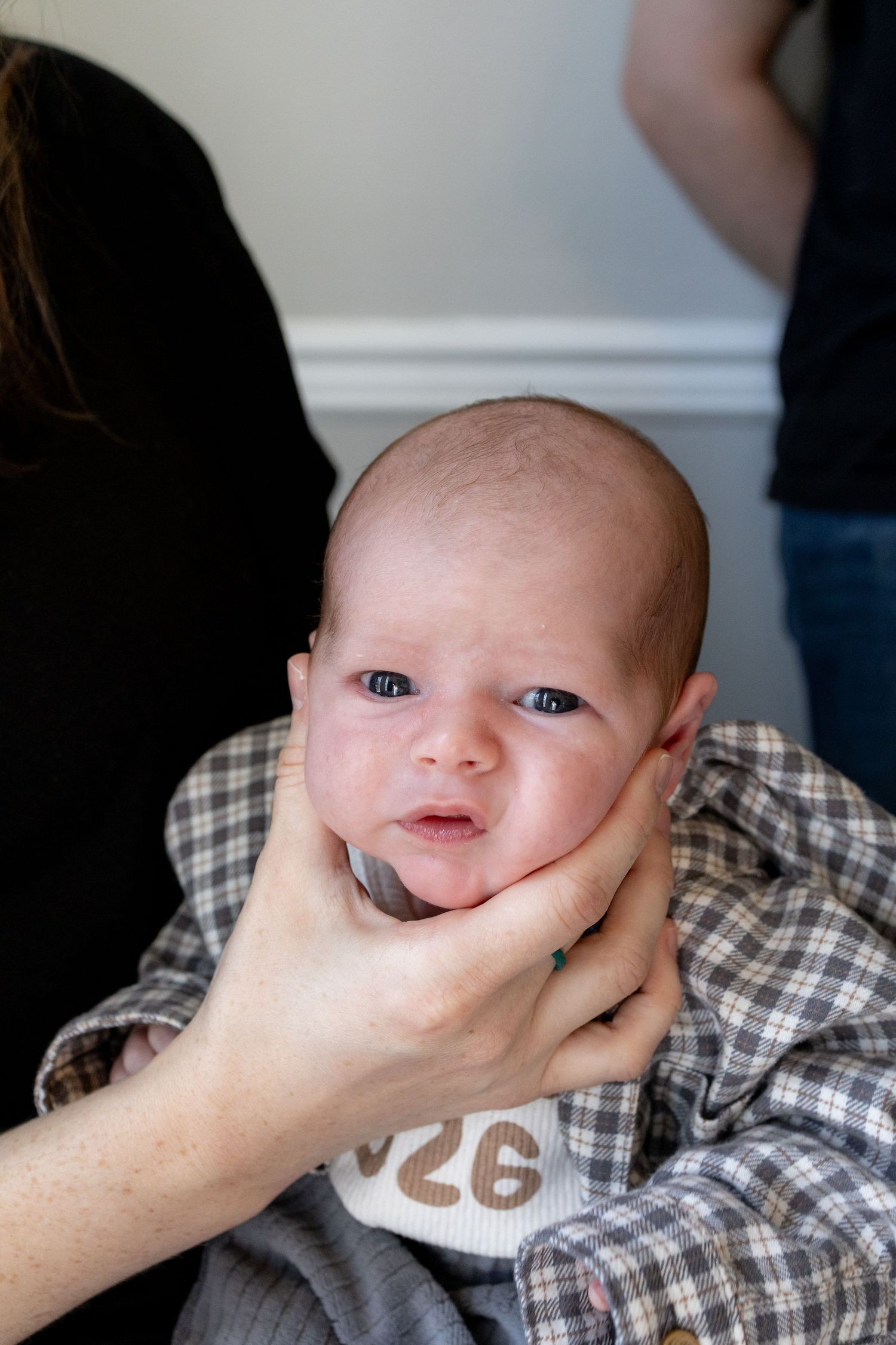 Newborn baby with blue eyes being held by adult hands, wearing plaid shirt and white bib.