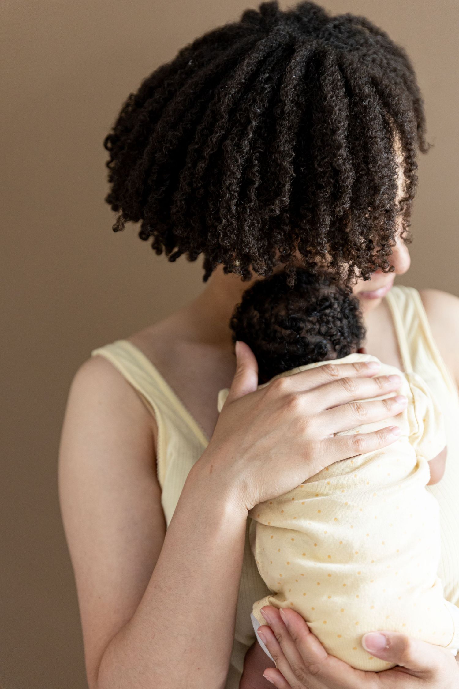 A mother with natural curly hair tenderly holds her newborn baby wrapped in a yellow swaddle blanket.