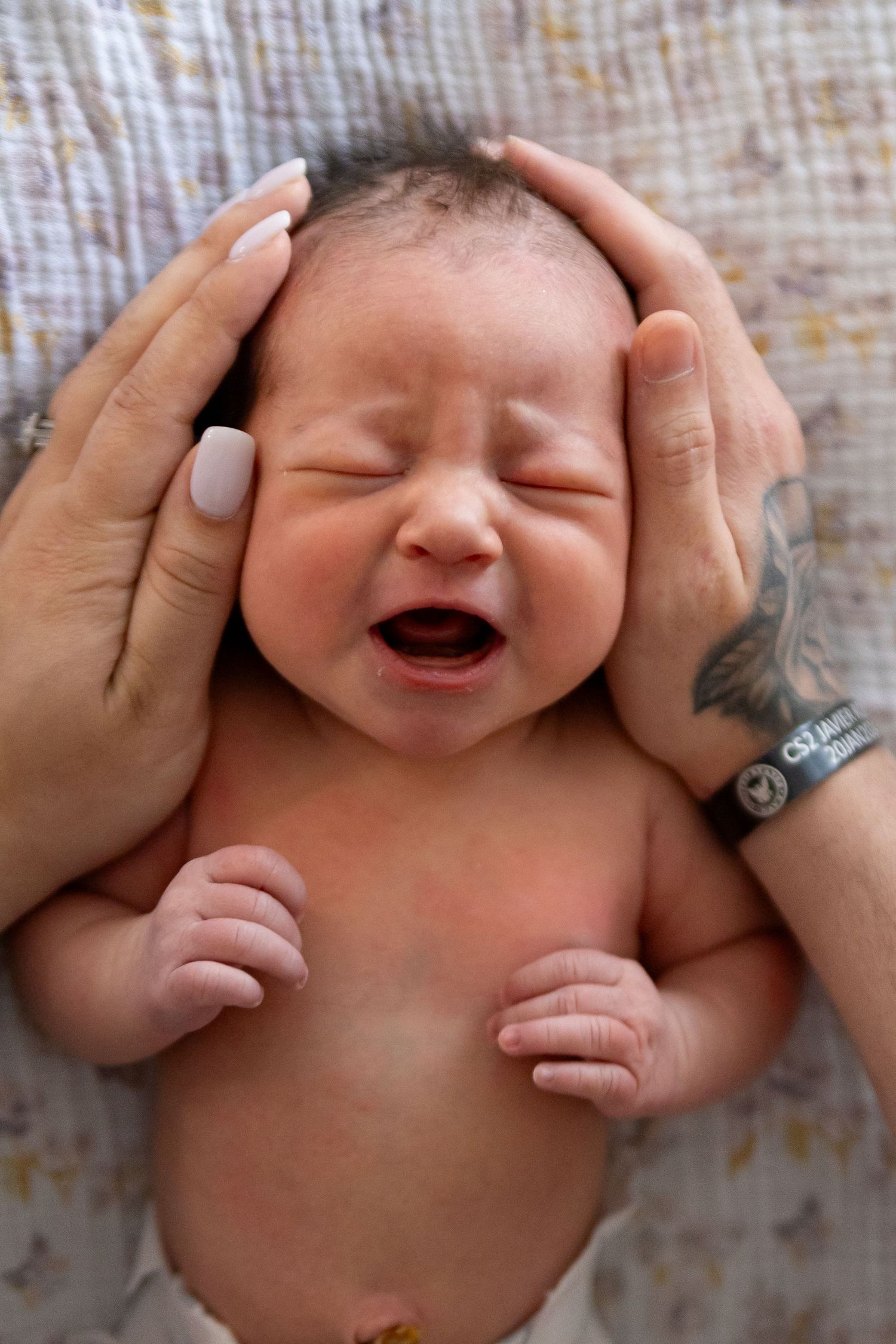 A newborn baby lies crying on soft fabric while gentle hands cradle their head from above.