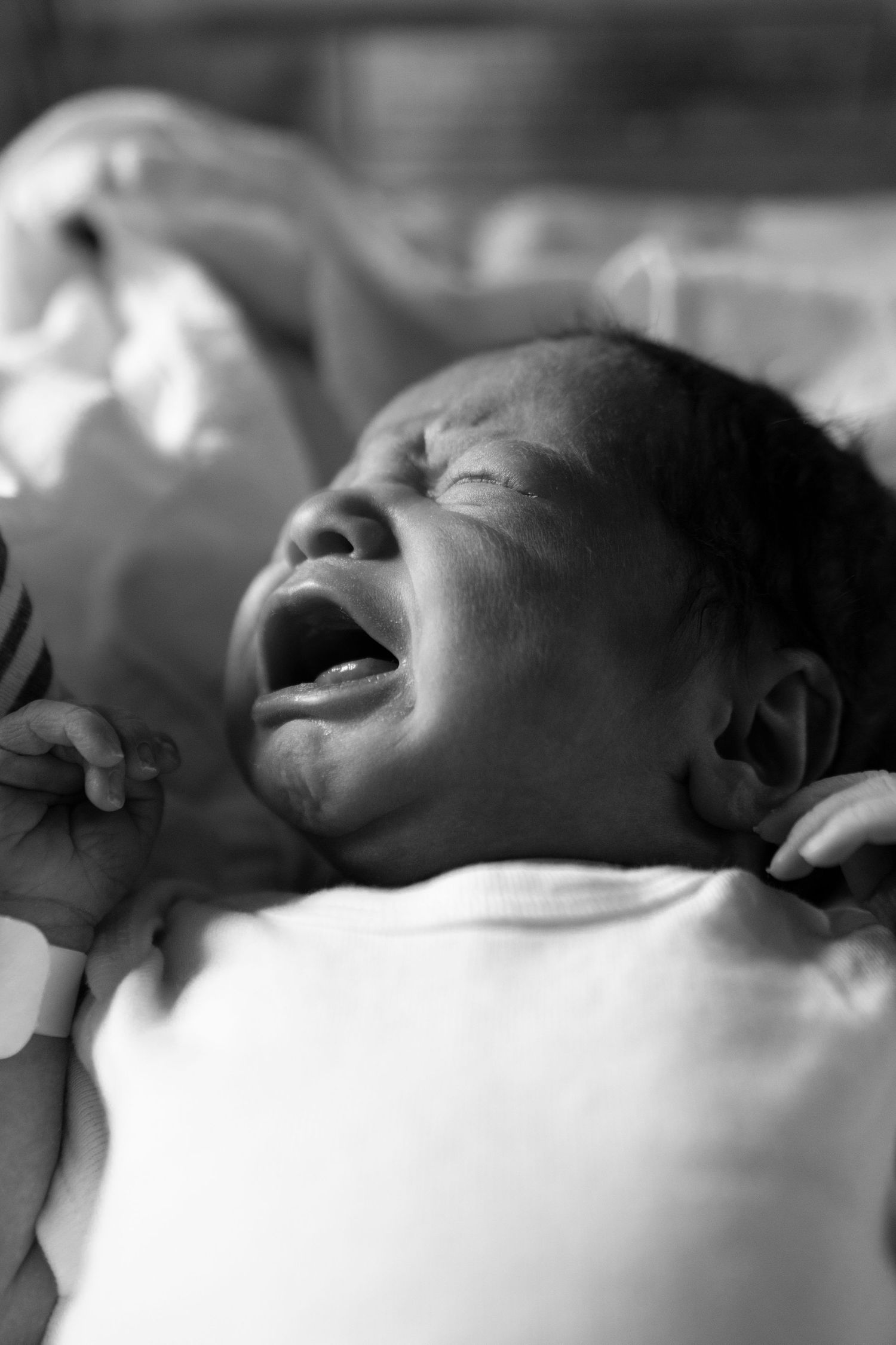 Black and white photo of a crying baby in white clothing laying down.