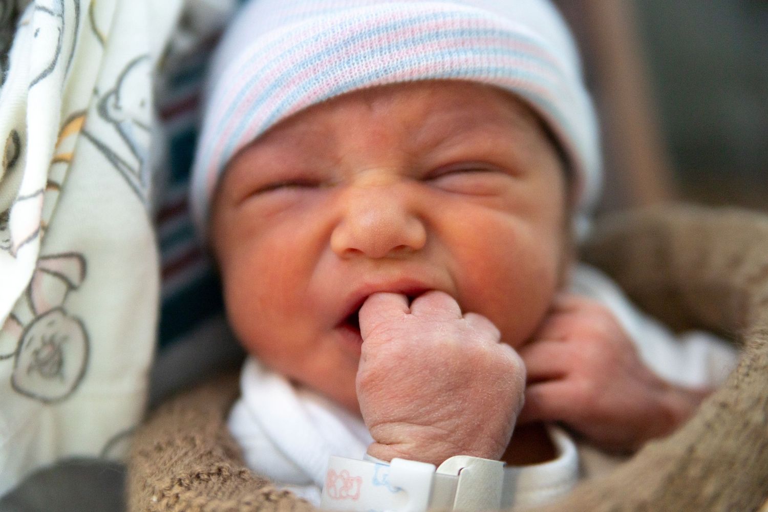 Newborn baby wearing white hat smiles with hand near mouth while resting on brown blanket.