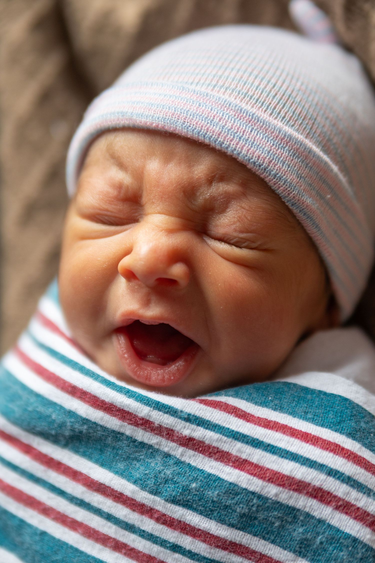 A newborn baby yawning while wrapped in a striped blanket and wearing a white knit hat.