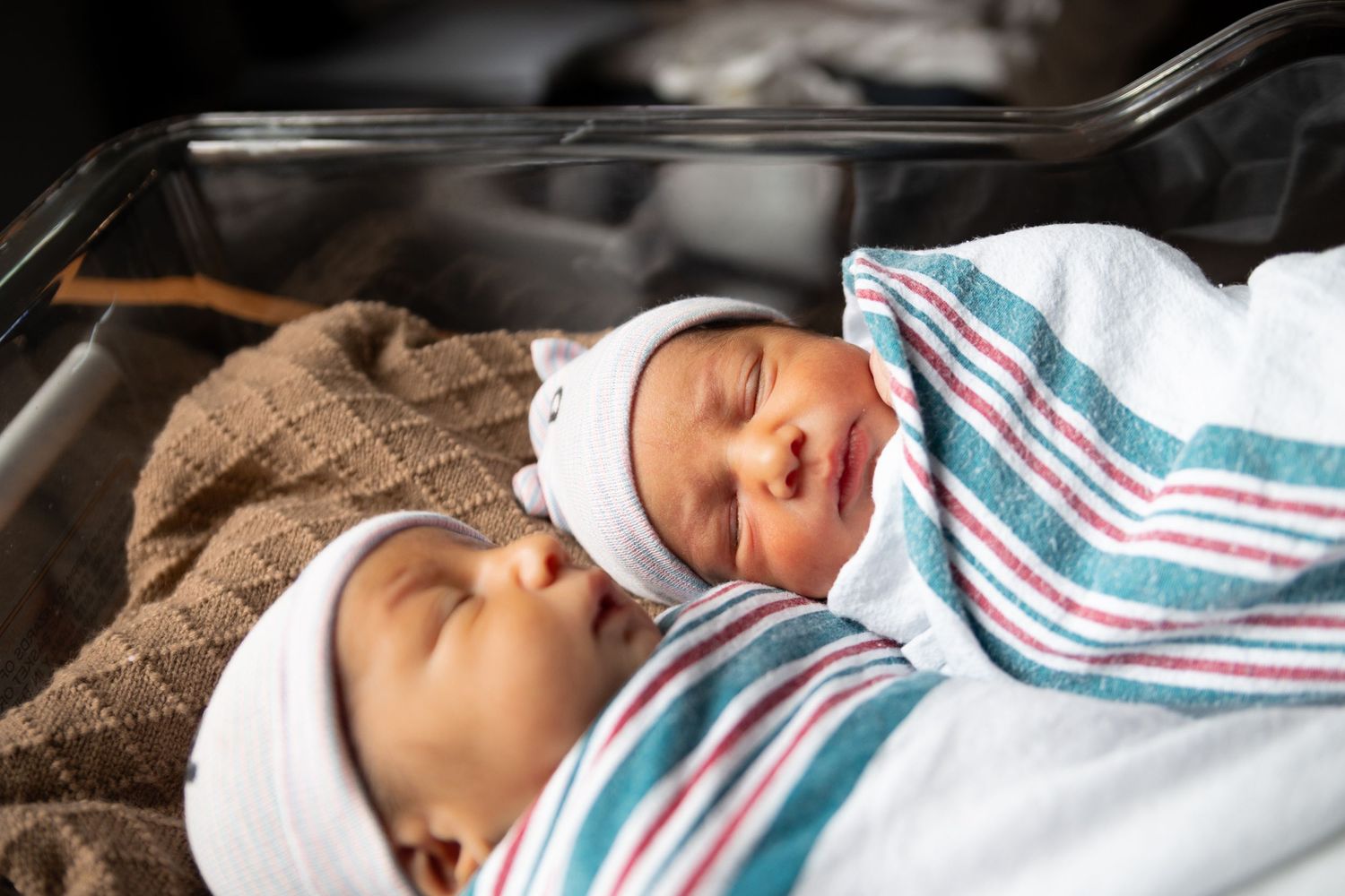 Two sleeping newborn babies snuggled together in white and teal striped hospital blankets.