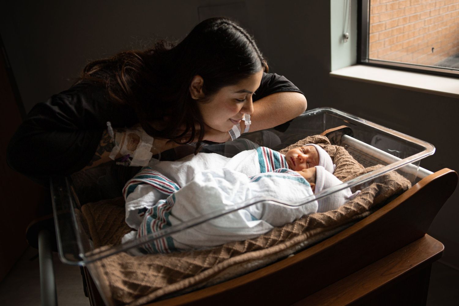 A mother gazes lovingly at her newborn baby sleeping in a hospital bassinet by a window.