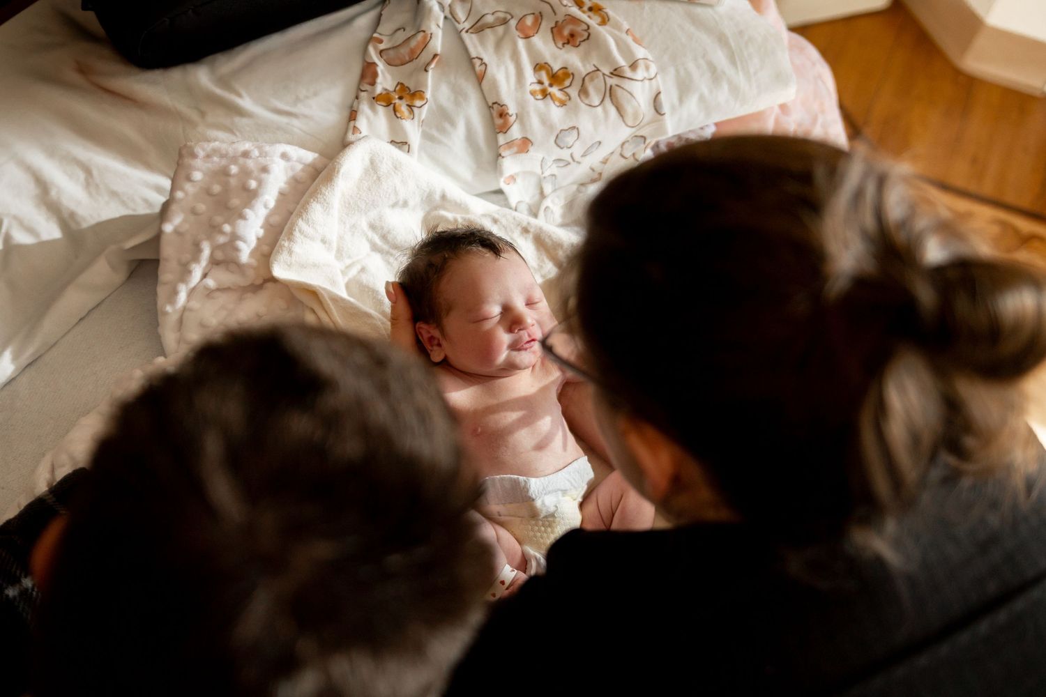 Several adults gather around a resting newborn baby on white bedding in warm lighting.