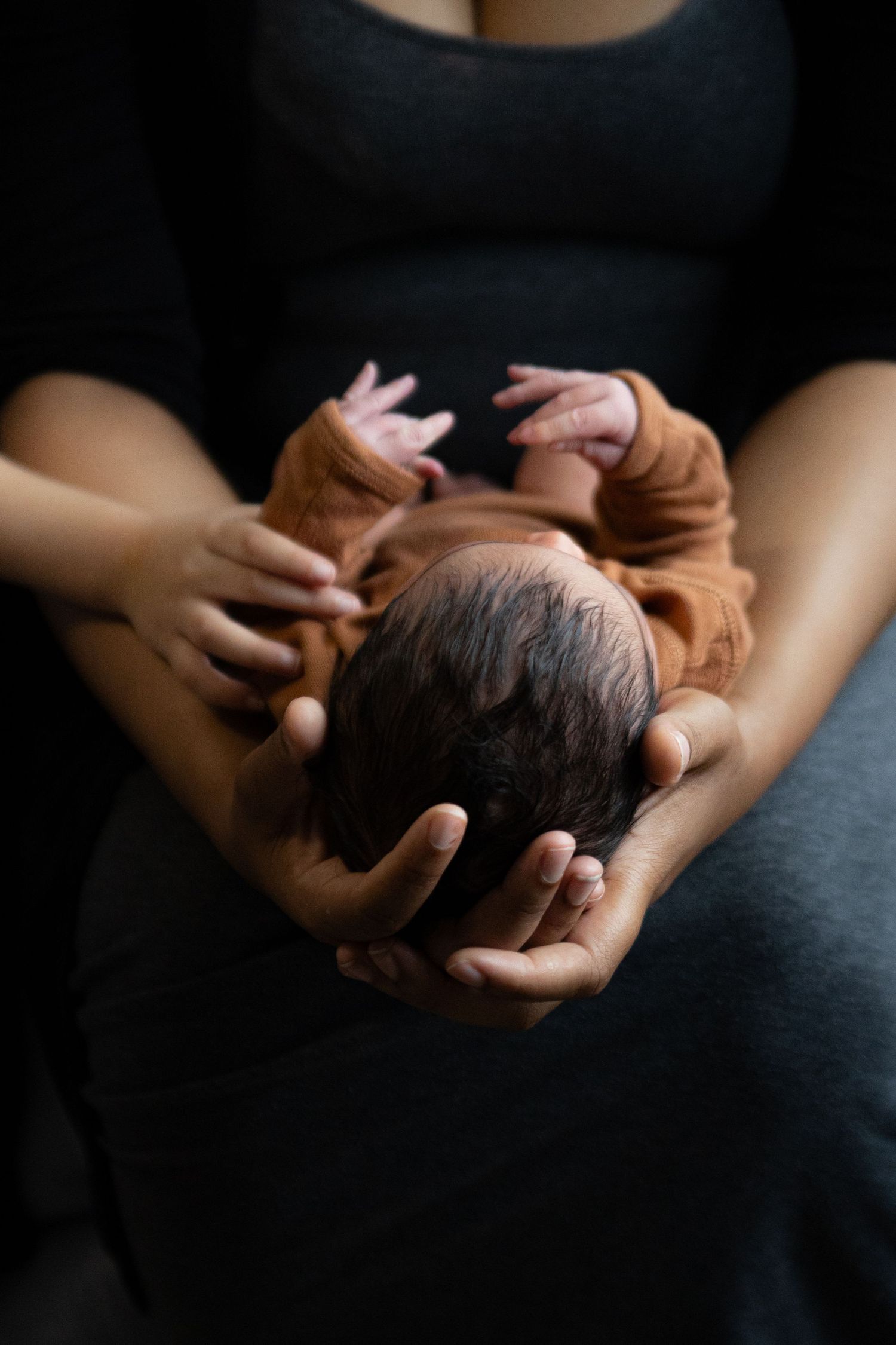 Gentle hands cradle a sleeping infant wearing a brown onesie against dark fabric in moody lighting.
