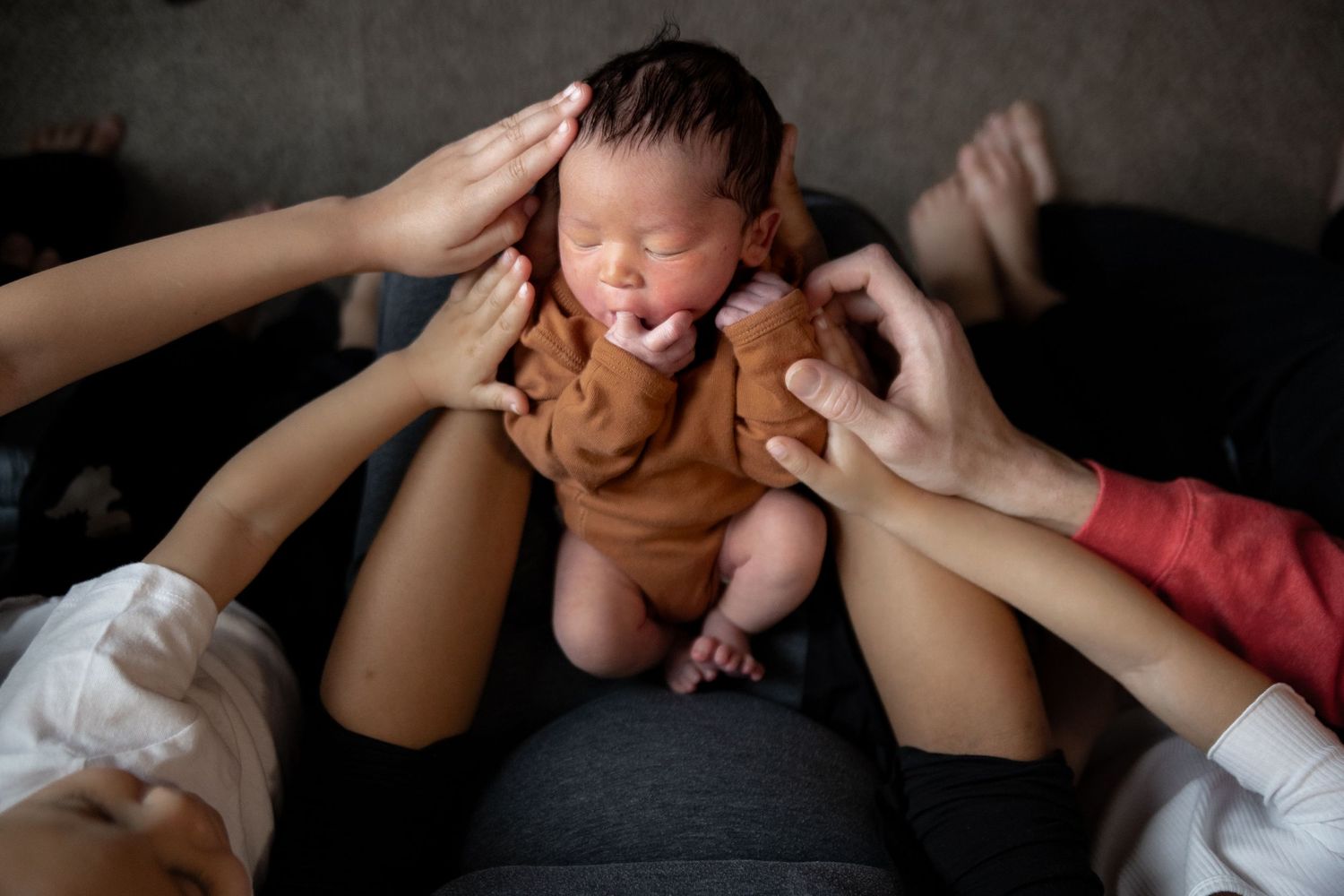 Multiple hands reach to hold and comfort a crying newborn baby in a brown onesie on a dark couch.