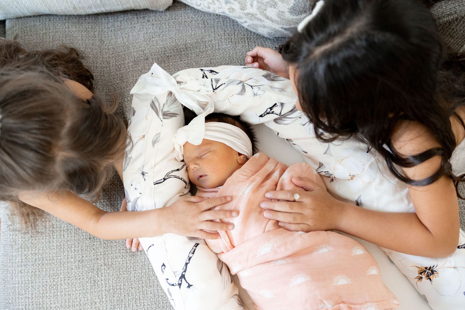 A newborn baby sleeps peacefully on patterned bedding while two people gently place their hands on the infant.