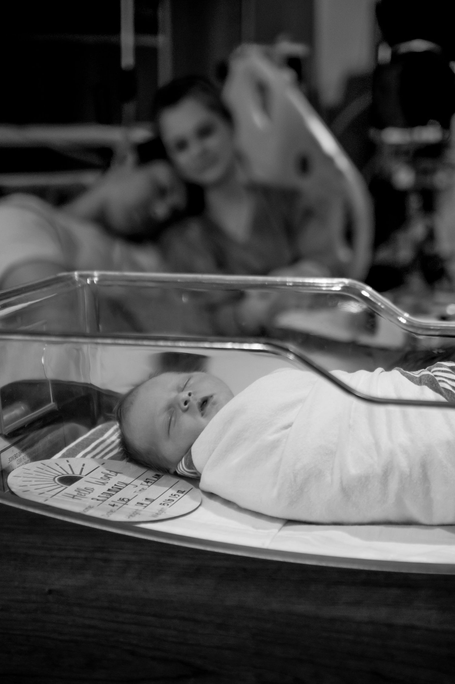 A newborn baby rests in a hospital bassinet while monitoring equipment is visible in the background.