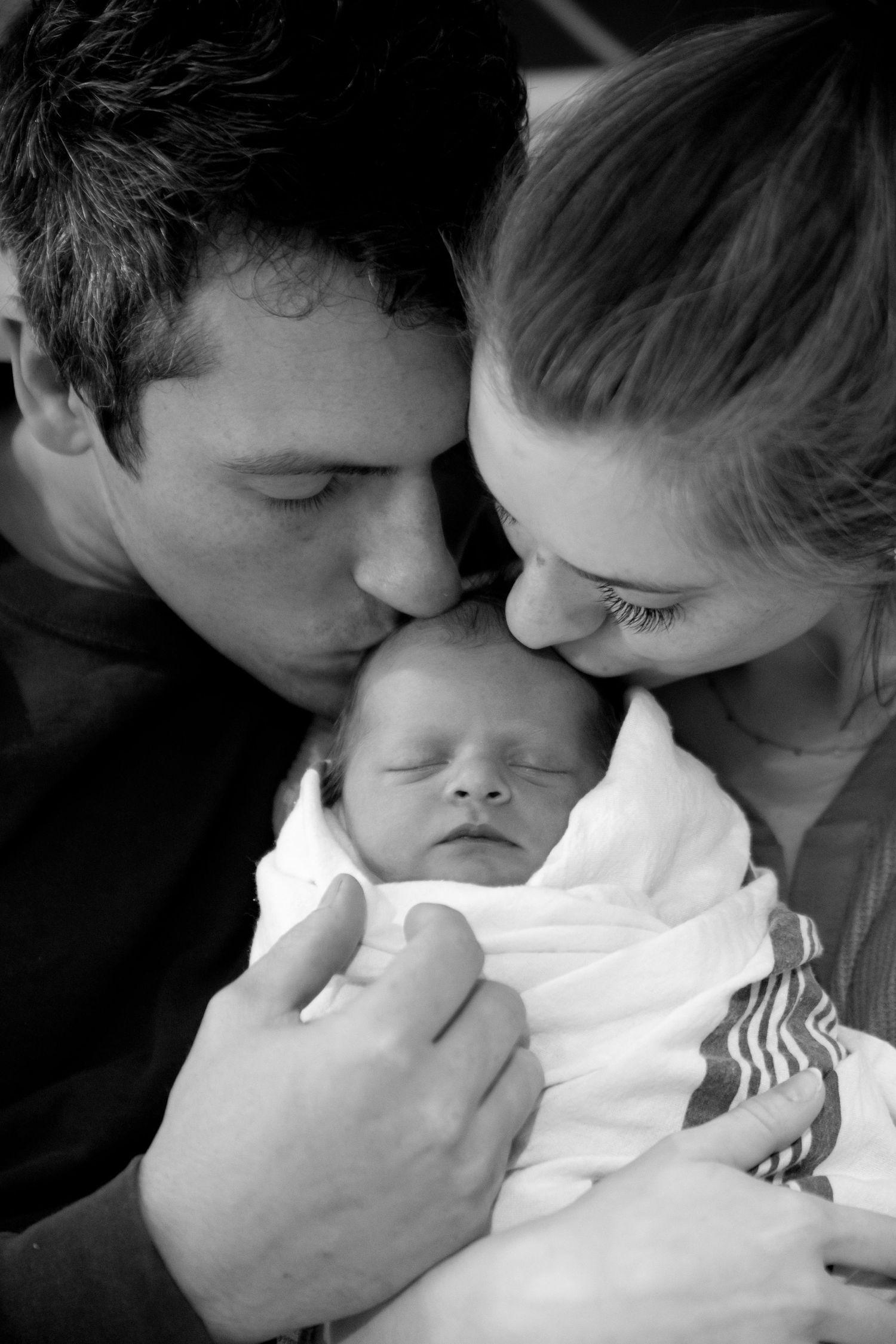 Intimate black and white portrait of parents sharing a tender moment with their sleeping newborn baby.