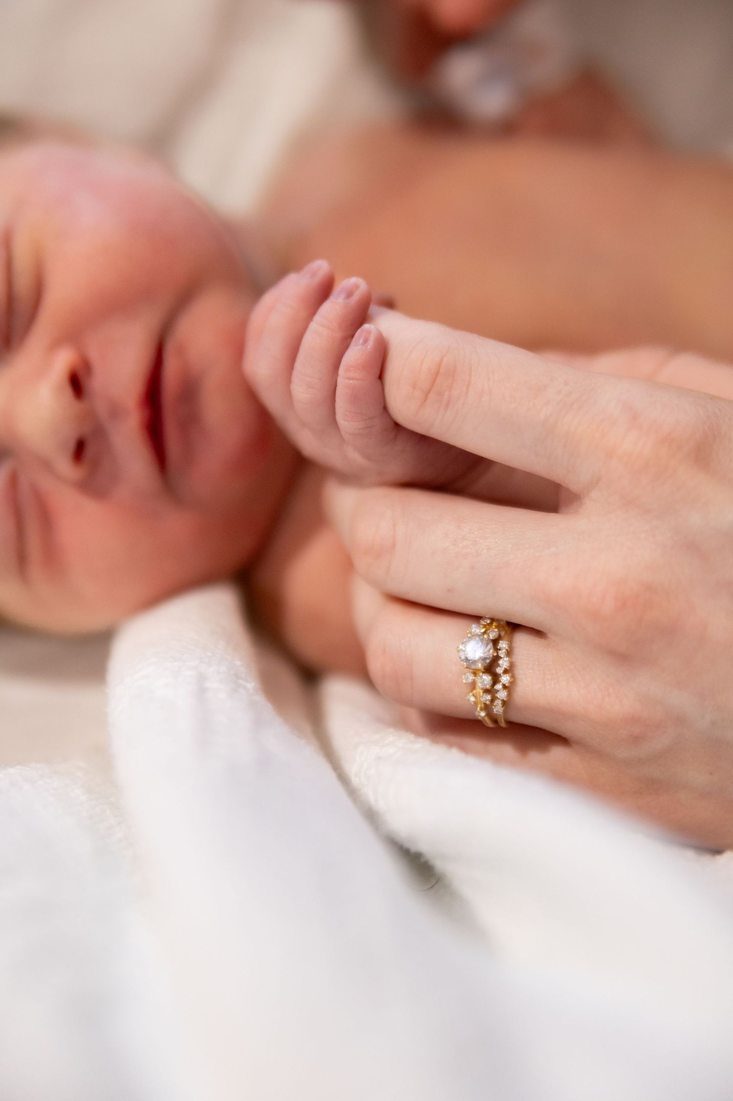 A tiny baby's hand grasps an adult finger wearing decorative gold rings against a soft white background.