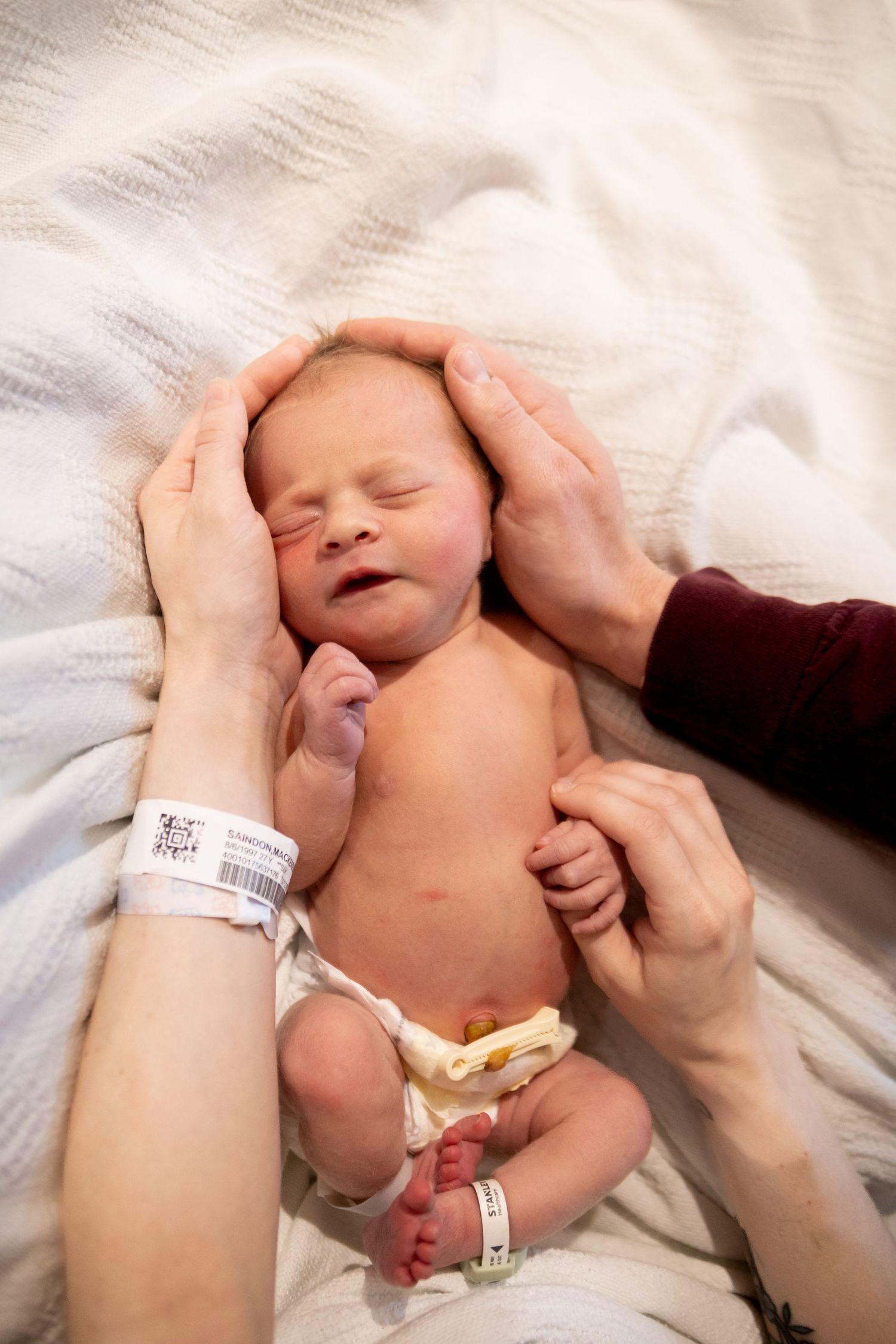 Newborn baby lying on white blanket with hospital ID bands while gentle hands cradle their head.