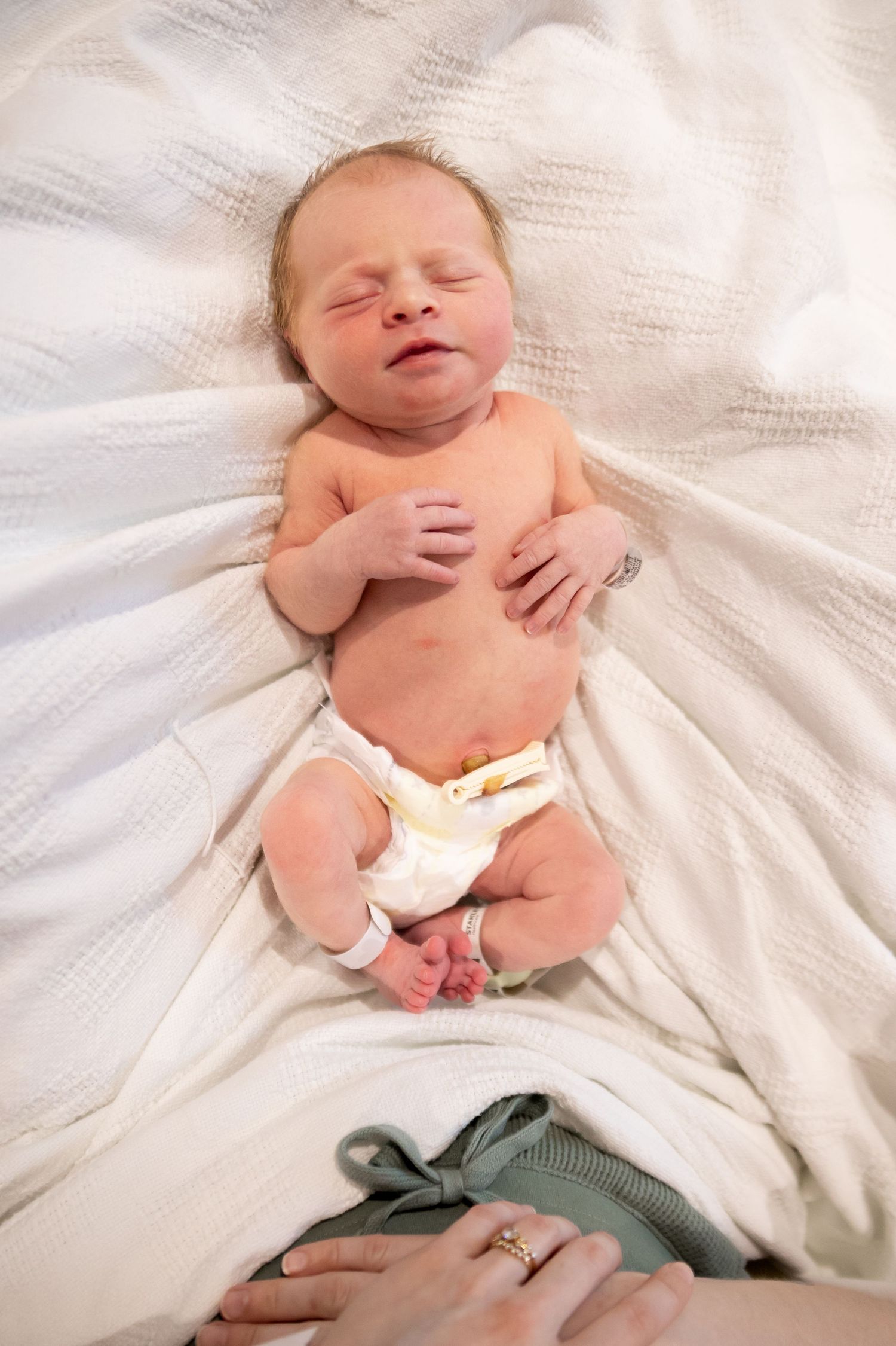 A peaceful sleeping newborn wearing a diaper lies on soft white fabric with an adult's hand visible at the bottom.