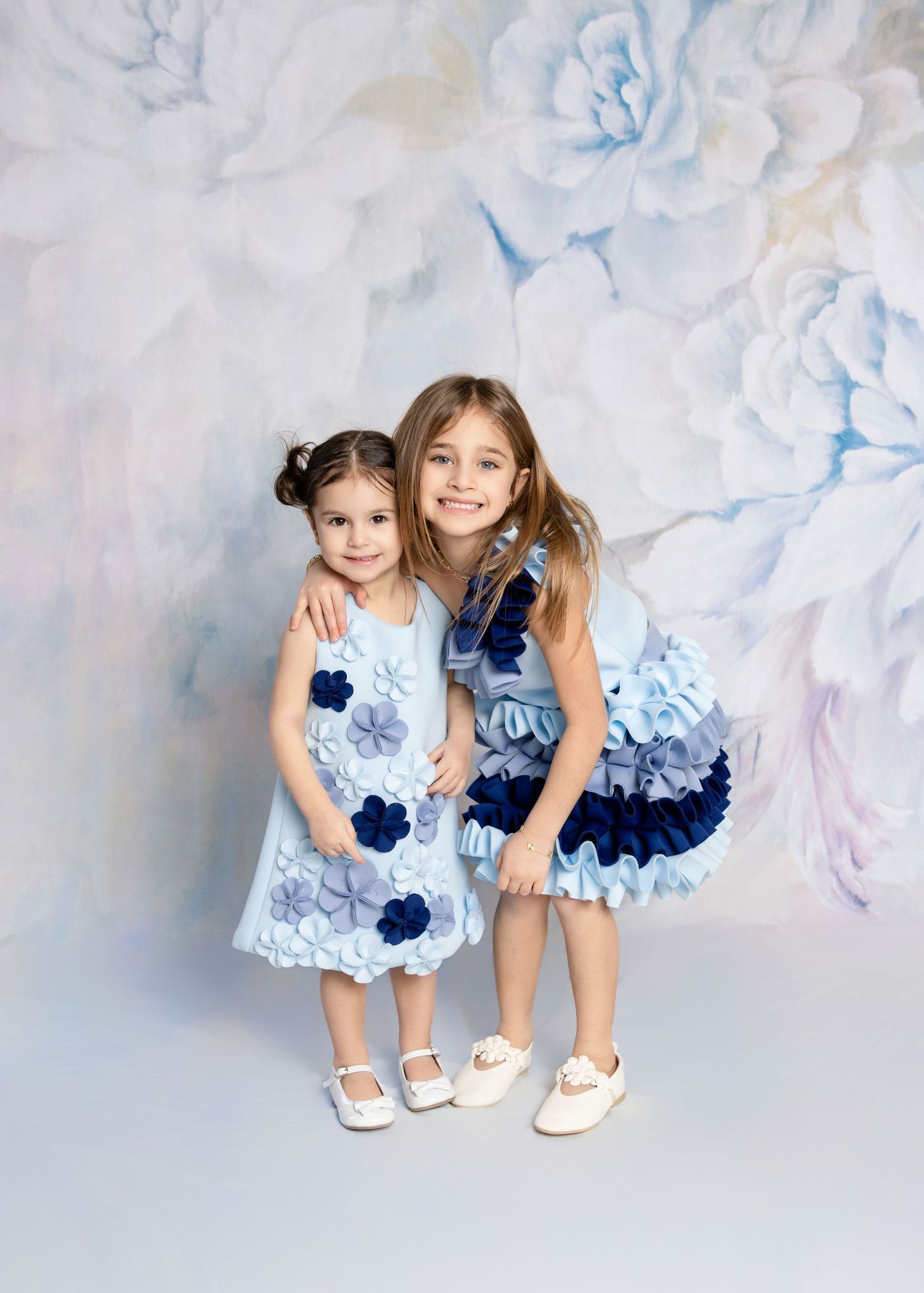 Two young girls in matching blue floral dresses pose together against a soft floral backdrop.